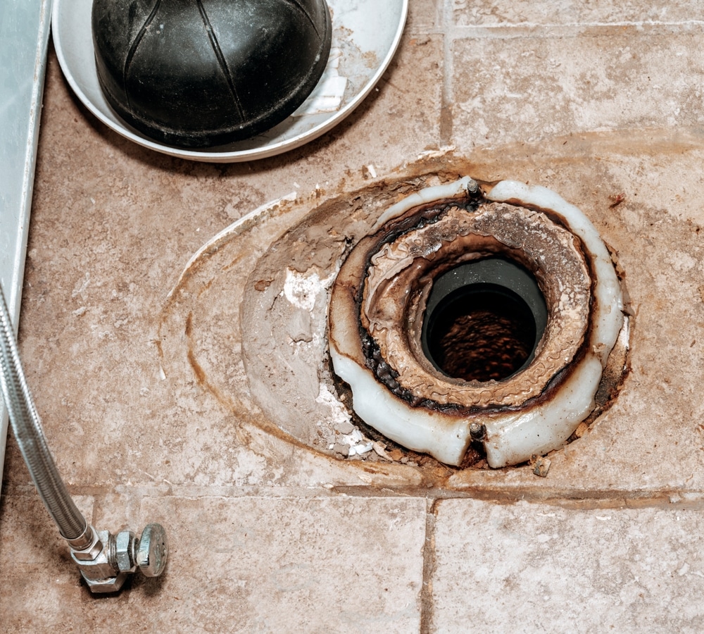 A close-up view of a toilet flange with a worn wax ring on a tiled bathroom floor in the house, next to plumbing fixtures and a plunger—potentially the cause of an unpleasant sewer odor.