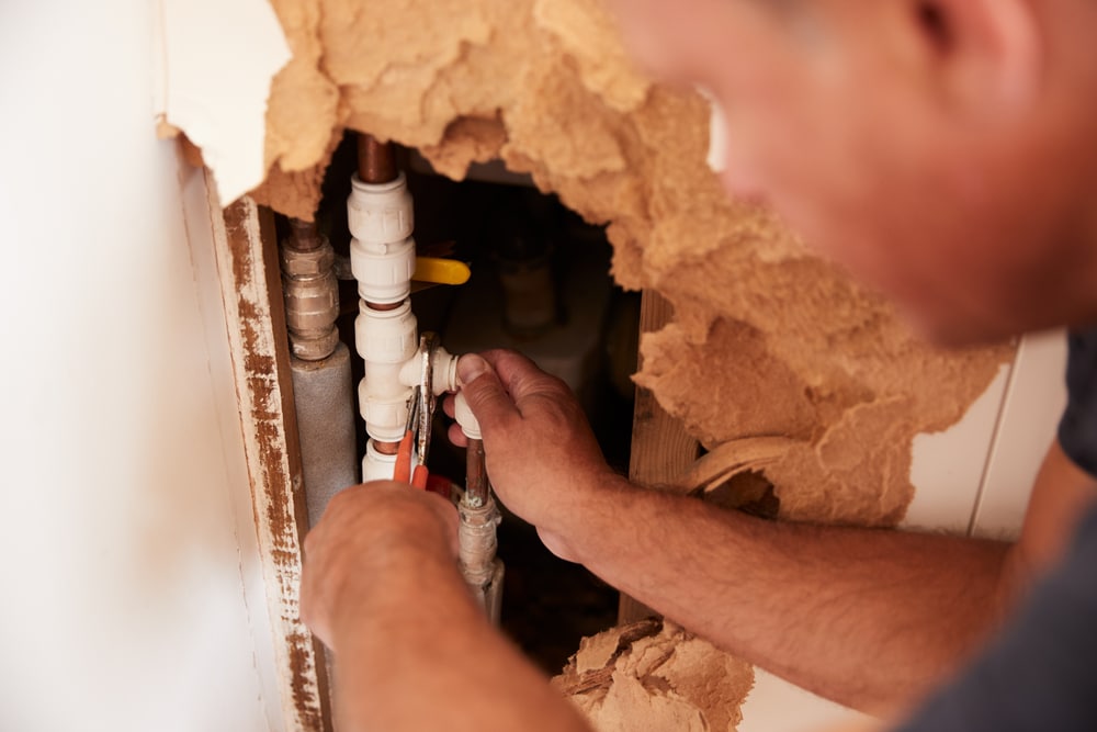 Person repairing plumbing inside a wall with damaged drywall, using tools to work on pipes behind the surface—addressing warning signs of burst pipes before further damage occurs.